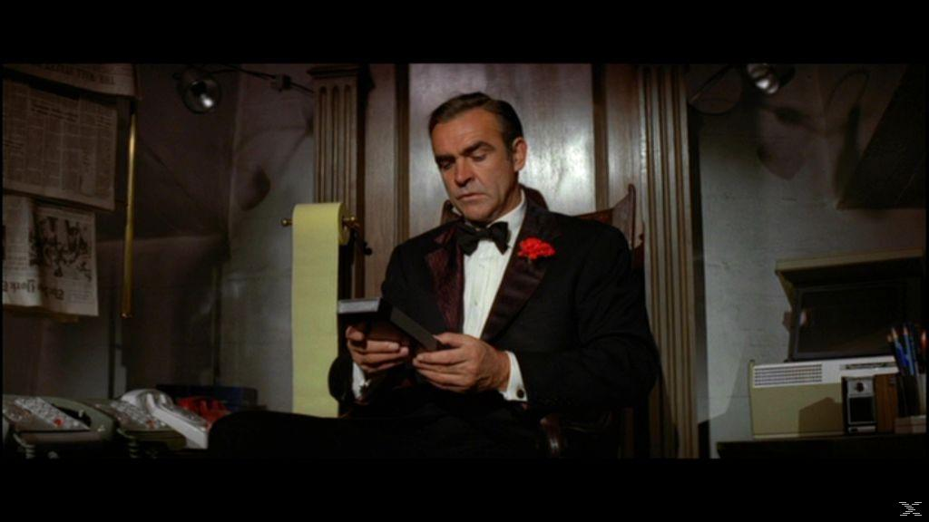 Sean Connery im Smoking betrachtet etwas. Eine rote Blume ist auf seiner Jacke. Eine Papierrolle und ein Telefon sind sichtbar.