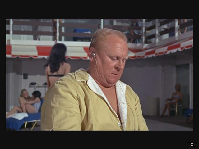 Ein Mann in einem gelben Bademantel mit Ohrhörer blickt nach unten. Frauen in Bikinis sind im Hintergrund in der Nähe eines Pools, einige sonnen sich.