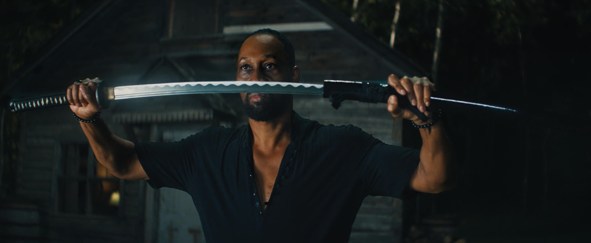 Ein Mann hält ein Katana horizontal. Er trägt ein schwarzes Hemd und steht vor einem Holzgebäude. Die Szene ist schwach beleuchtet.