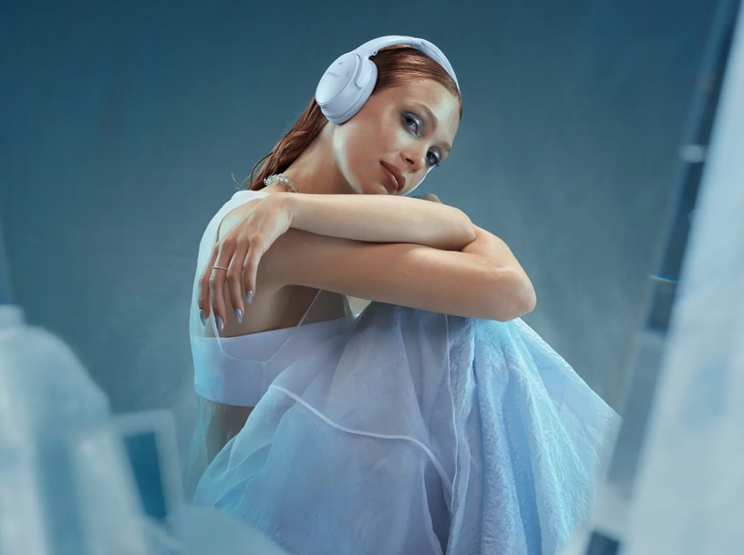 Eine Frau mit roten Haaren, die einen weißen Kopfhörer und ein hellblaues Kleid trägt, sitzt. Sie schaut zur Kamera.