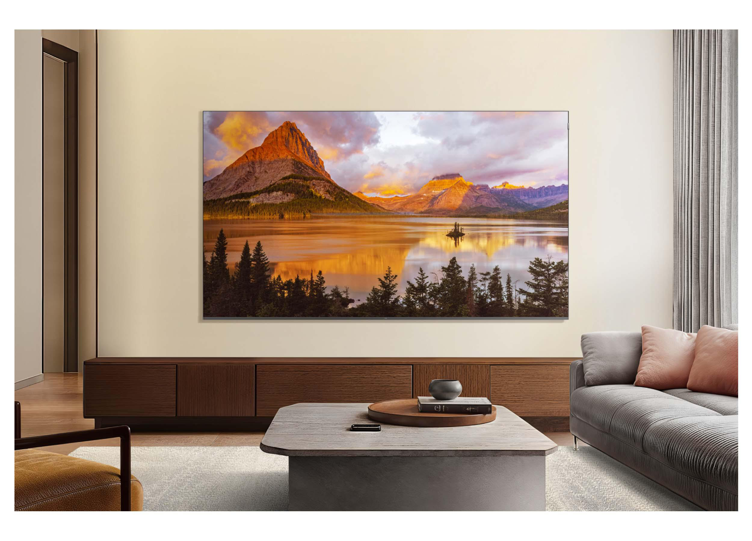 Wohnzimmer mit Fernseher, der eine Bergseeszene zeigt, Holzmöbeln, grauem Sofa und einem Couchtisch.