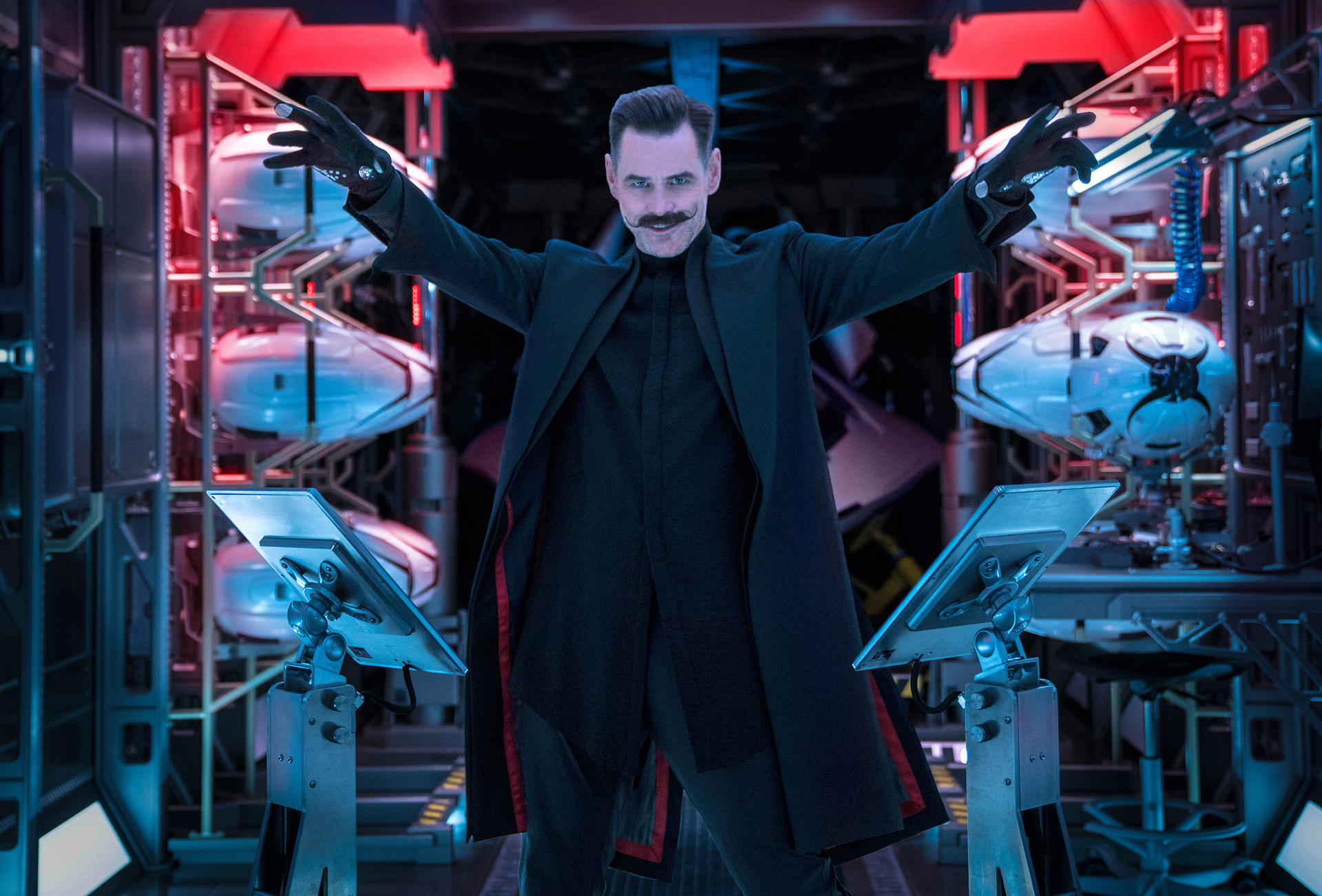 Jim Carrey als Dr. Robotnik posiert mit ausgebreiteten Armen. Im Hintergrund befinden sich rot und blau beleuchtete Laborgeräte. Er trägt Schwarz.