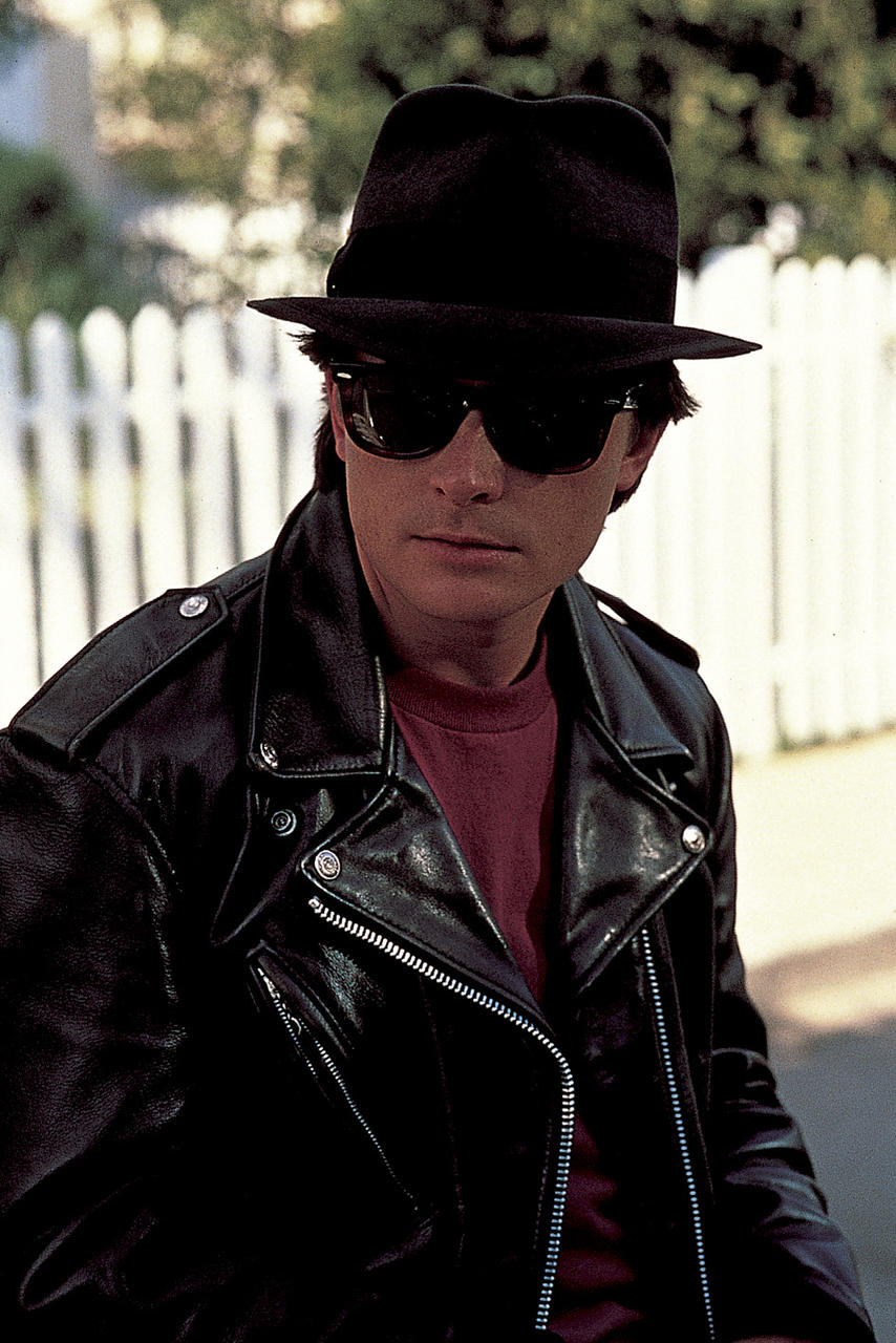 Mann mit schwarzem Hut und Sonnenbrille, Lederjacke und rotem Hemd, mit weißem Zaun im Hintergrund.
