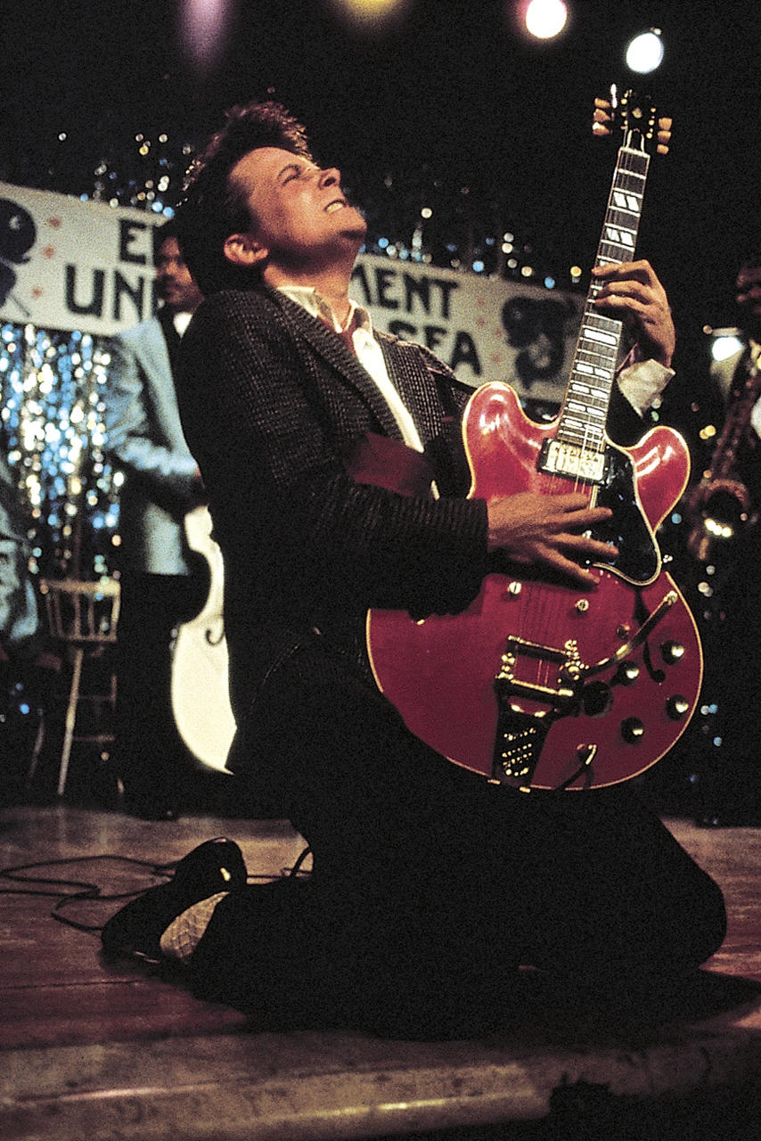 Ein Mann kniet auf der Bühne und spielt eine rote Gitarre. Er trägt einen Anzug und blickt nach oben. Im Hintergrund sind ein Banner und Bandmitglieder zu sehen.