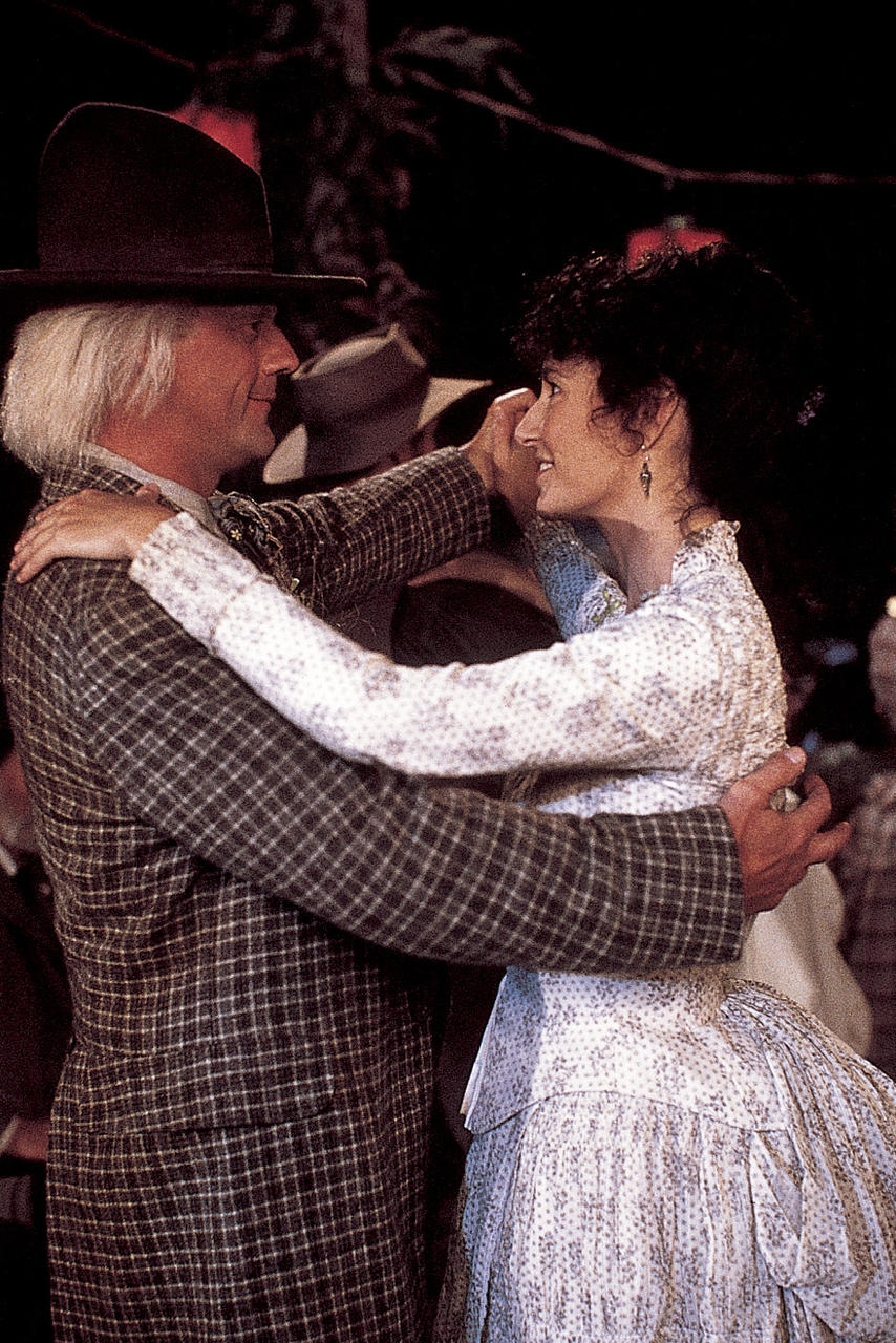 Ein Mann in karierter Jacke und Hut tanzt mit einer Frau in floralem Kleid. Sie umarmen sich und lächeln. Im Hintergrund ist eine Menschenmenge zu sehen.