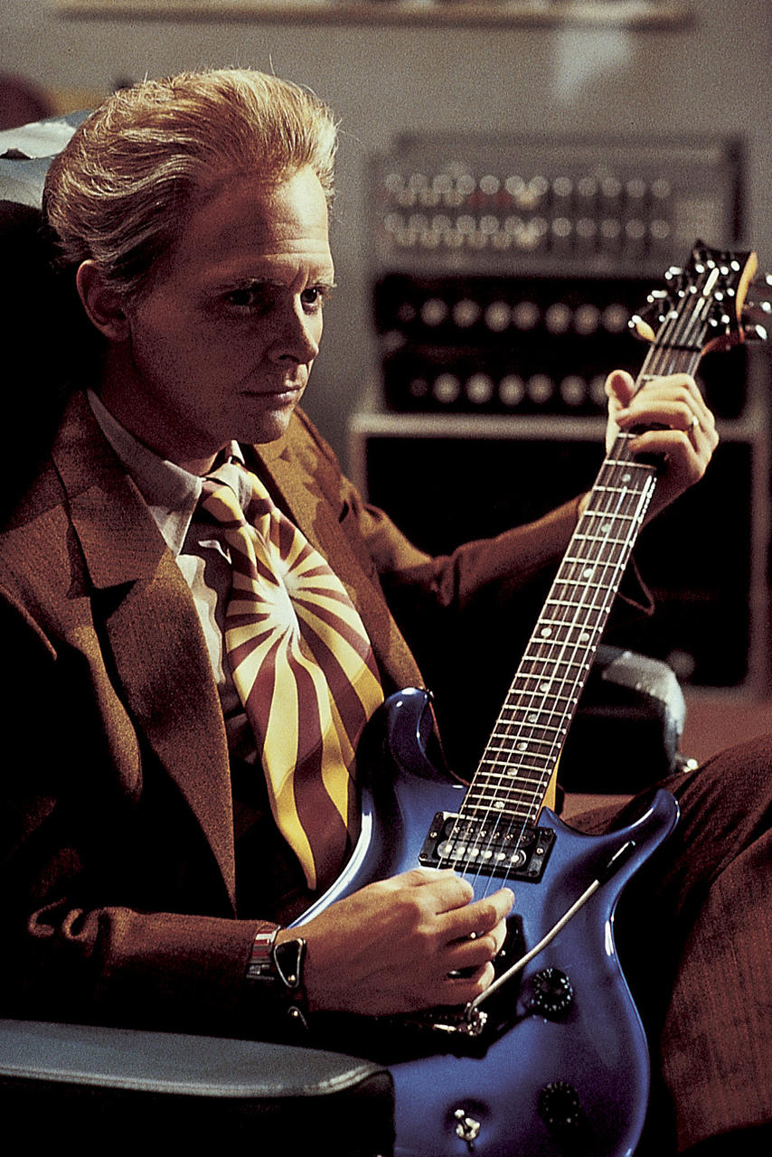 Mann mit blauer Gitarre. Er trägt einen braunen Anzug und eine gemusterte Krawatte. Im Hintergrund Studioausrüstung.