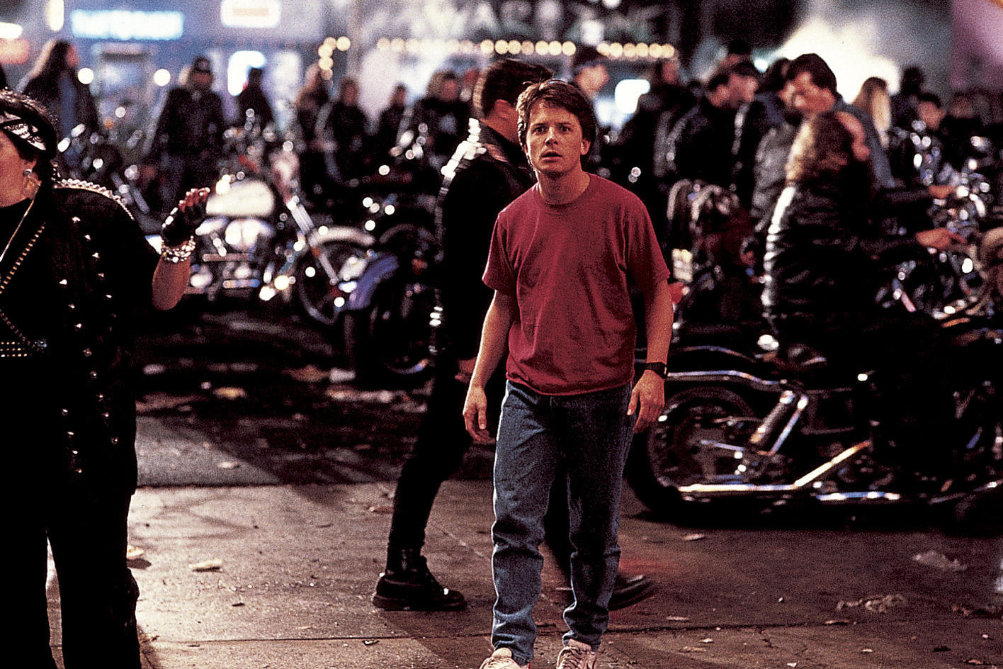 Ein Mann in rotem Hemd und Jeans geht auf den Betrachter zu. Im Hintergrund sind Motorräder und Menschen bei Nacht.