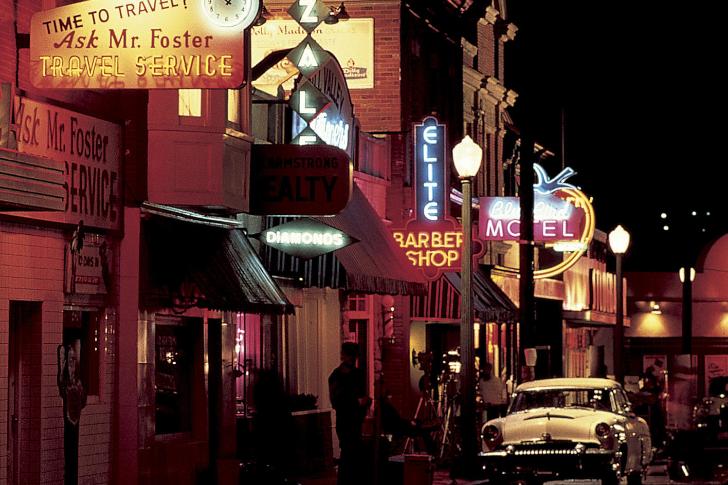 Eine Nachtszene mit Neonreklamen für Reisebüro, Immobilien, Friseur und Motel. Ein klassisches Auto steht auf der Straße.