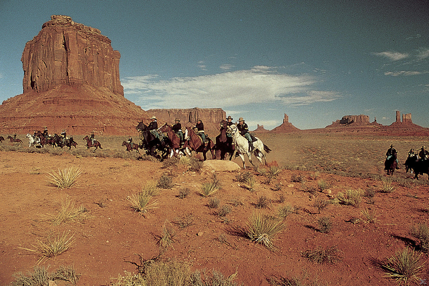Cowboys auf Pferden reiten durch eine Wüstenlandschaft. Rote Felsformationen und blauer Himmel im Hintergrund.