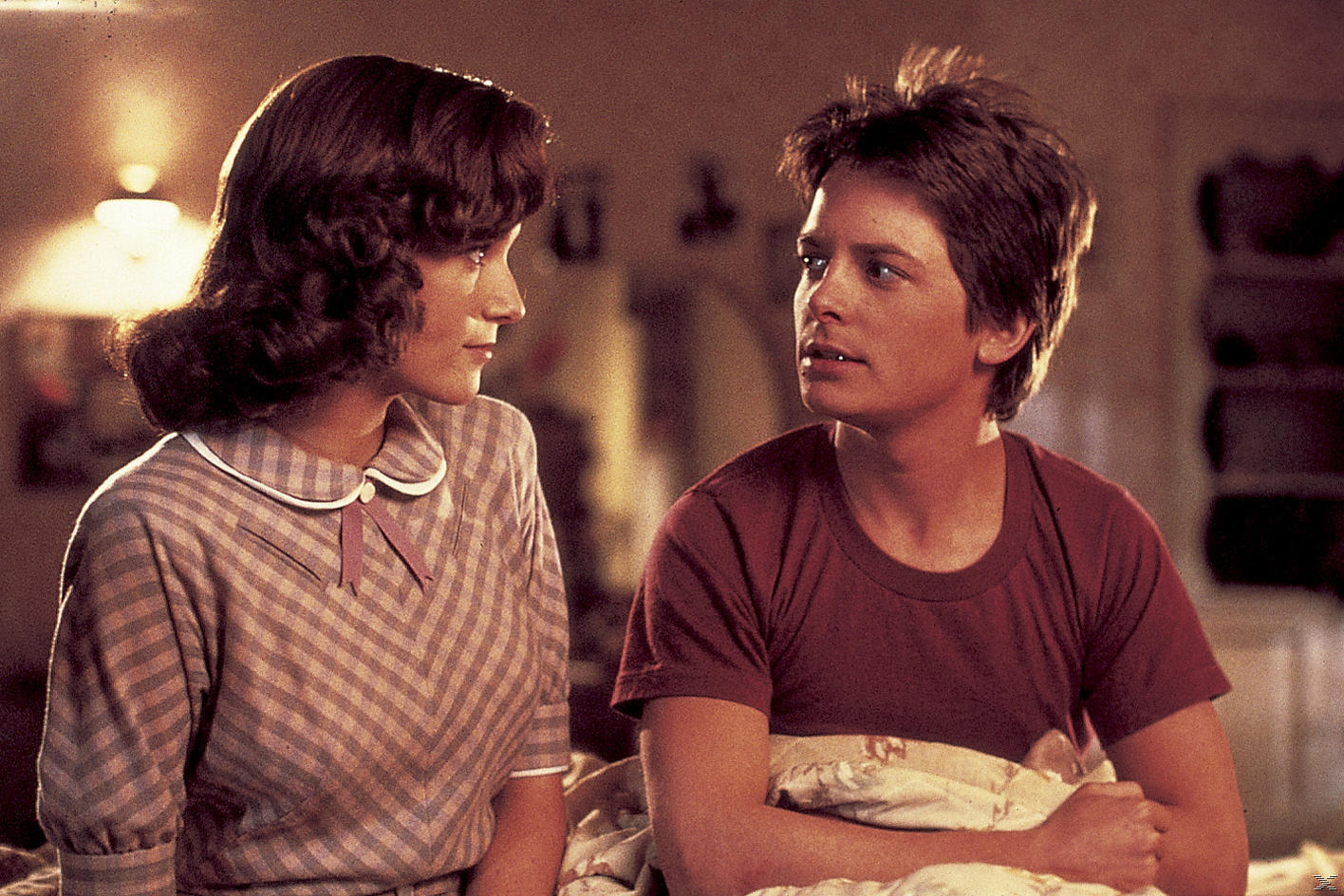 Ein Mann und eine Frau stehen sich gegenüber. Der Mann trägt ein rotes Hemd, die Frau ein gemustertes Kleid. Weiches Licht deutet auf eine Schlafzene hin.