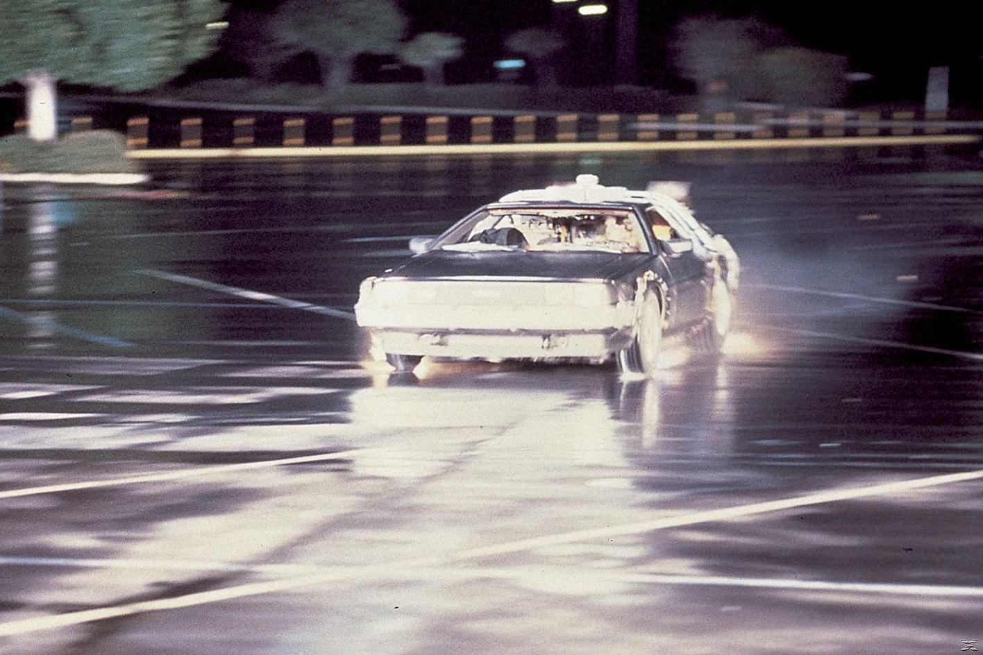 Ein DeLorean-Auto fährt nachts auf einer nassen Straße. Lichter spiegeln sich auf der Straße. Bäume und Gebäude sind im verschwommenen Hintergrund.