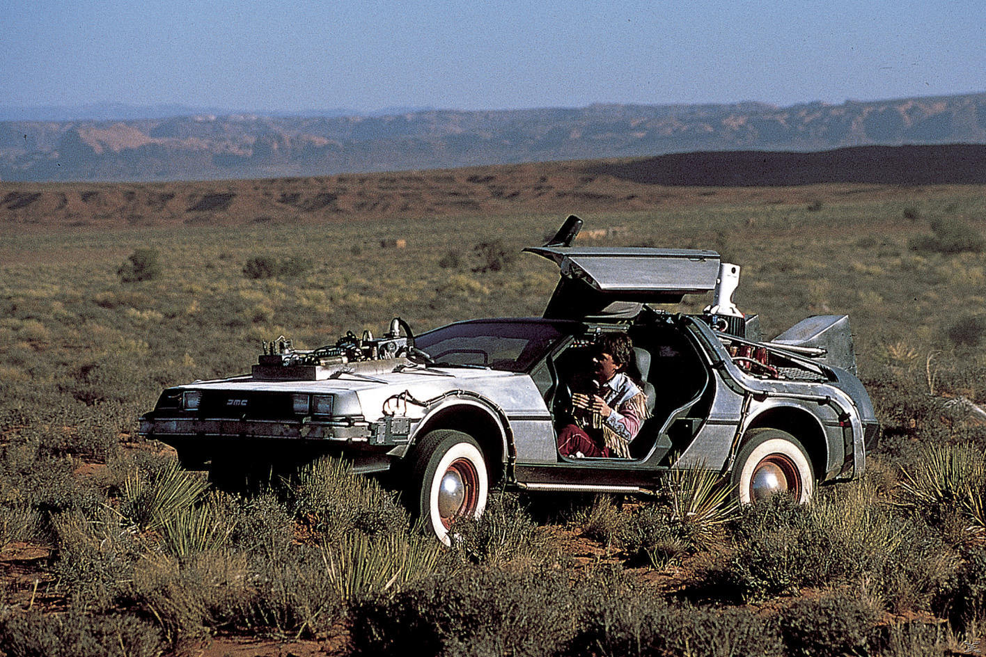 Eine Zeitmaschine vom Typ DeLorean steht in einer Wüstenlandschaft. Eine Person sitzt darin. Hügel sind im Hintergrund. Die Autotüren sind geöffnet.