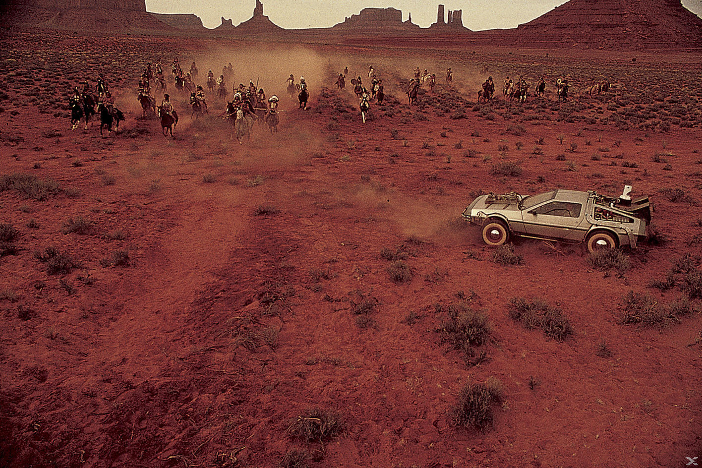DeLorean-Auto auf roter Erde mit Reitern auf Pferden. Staub steigt auf. Berge im Hintergrund, Himmel bedeckt.