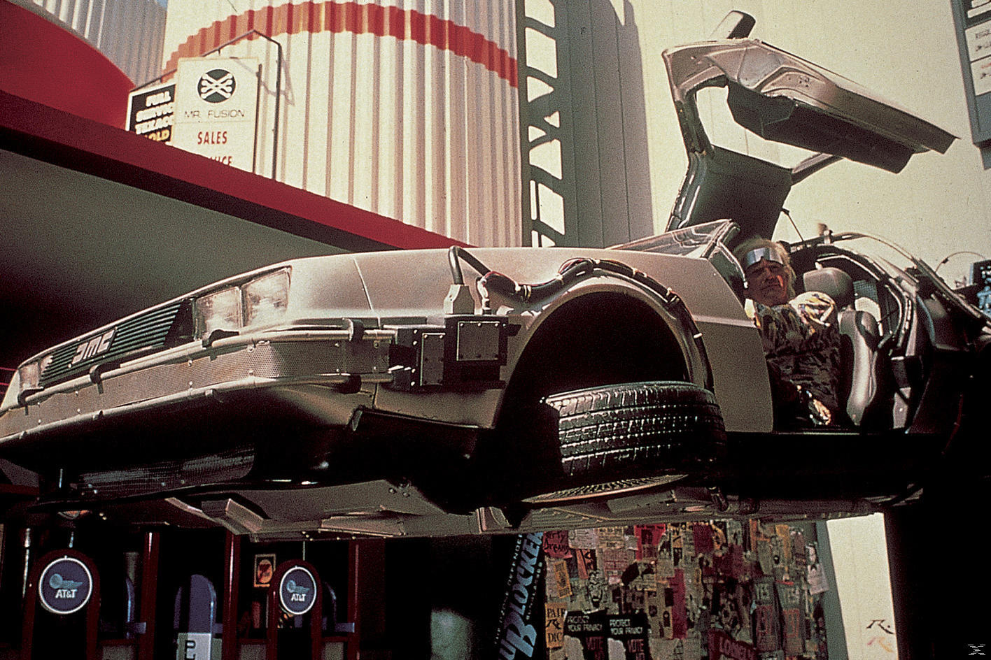 Delorean Zeitmaschine mit offener Tür. Mann am Steuer. Beschilderung im Hintergrund, AT&T-Logo unten.