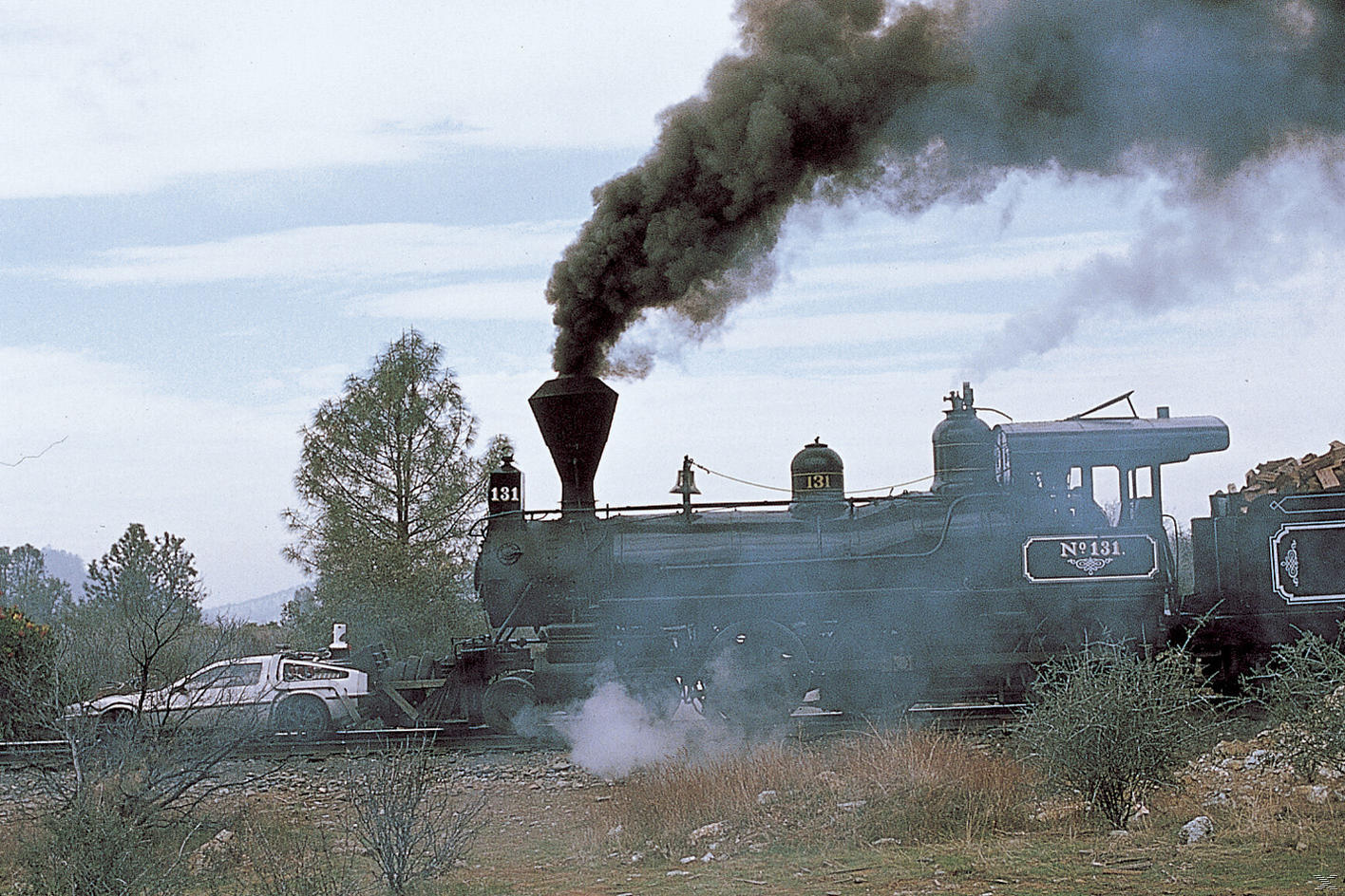 Eine Dampflok mit der Nummer 131 stößt schwarzen Rauch aus. Ein DeLorean-Auto steht neben dem Zug auf einem Gleis in einer Graslandschaft.