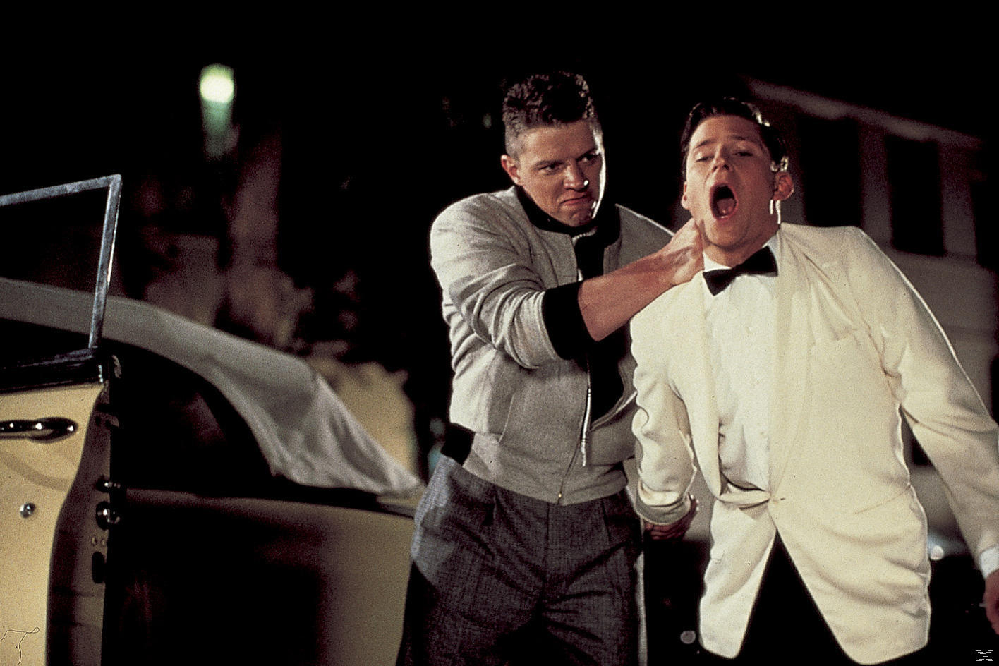 Ein Mann würgt einen anderen in einem weißen Anzug. Der Angreifer trägt eine graue Jacke. Links ist ein Auto zu sehen.