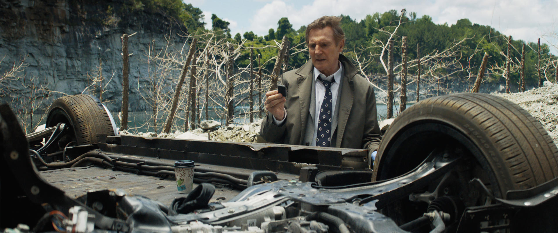 Liam Neeson in einem Mantel, der auf ein Telefon blickt und in der Nähe eines Fahrzeugunterbodens steht. Hintergrund: ein Steinbruch mit Bäumen.