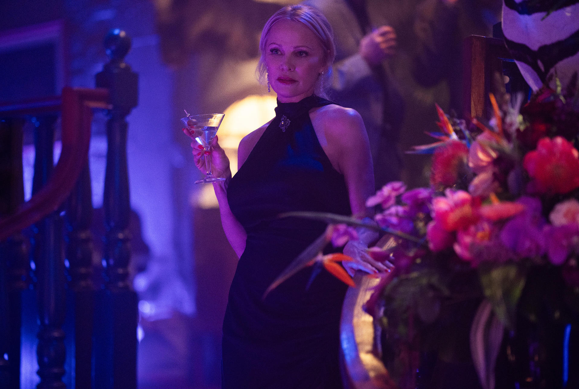 Blonde Frau in schwarzem Kleid hält Martini. Blumen im Vordergrund und Treppe im Hintergrund.