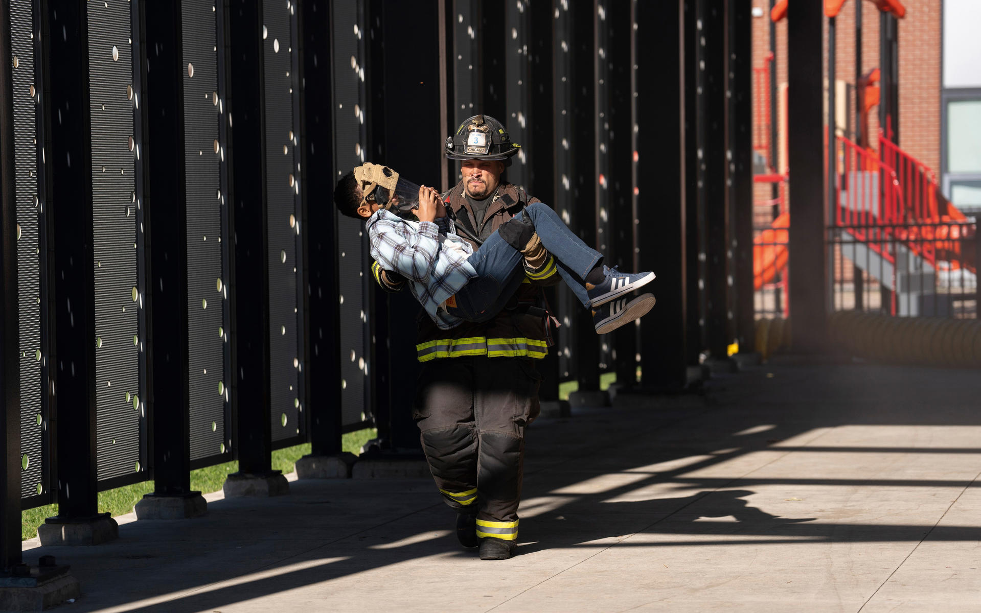 Ein Feuerwehrmann trägt ein Kind mit Maske. Sie gehen an einem Zaun vorbei. Im Hintergrund ist ein Spielplatz.
