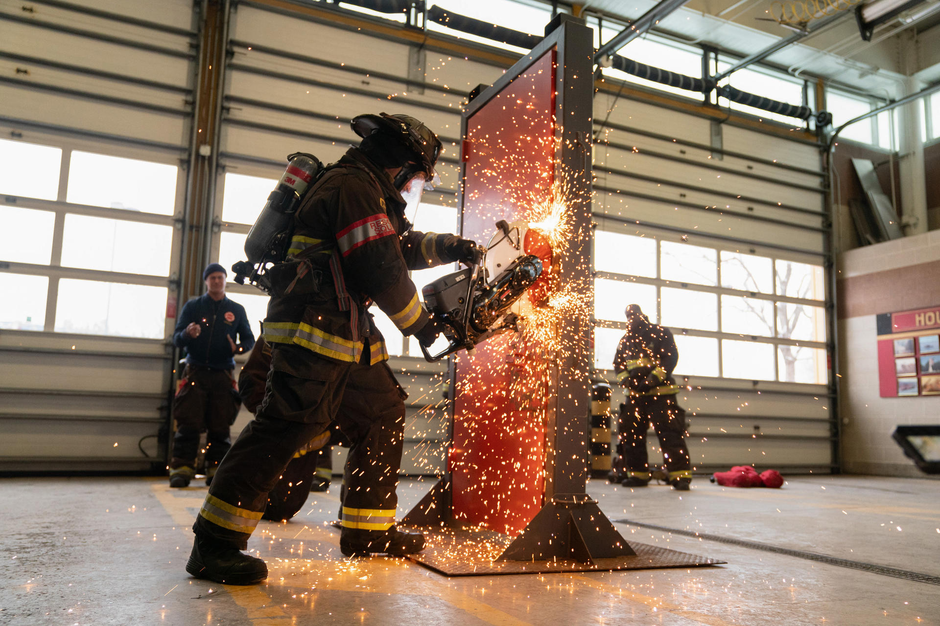Ein Feuerwehrmann schneidet eine Metallbarriere durch, Funken fliegen. Andere Feuerwehrleute und ein Garagentor sind im Hintergrund.