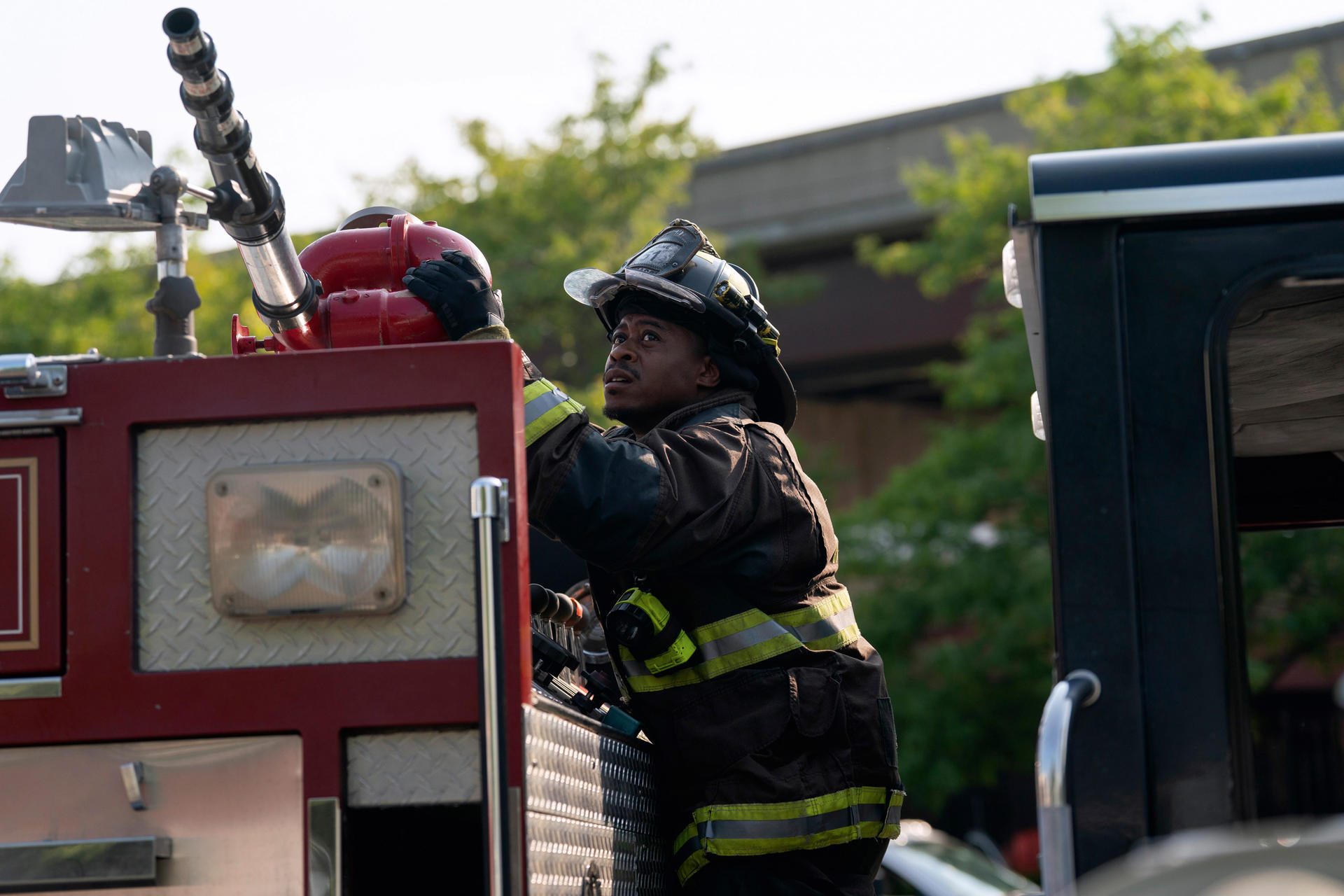 Ein Feuerwehrmann in Uniform auf einem Feuerwehrwagen. Er blickt nach oben und hält eine Wasserpumpe. Bäume und Gebäude im Hintergrund.
