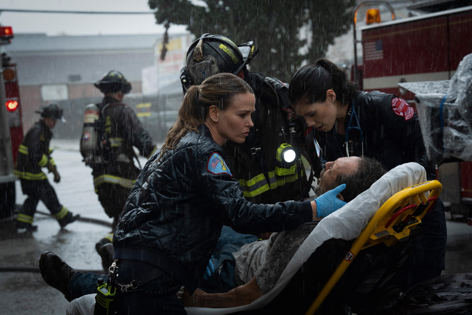 Feuerwehrleute und Sanitäter kümmern sich im Regen um einen Mann auf einer Trage. Feuerwehrwagen stehen im Hintergrund.