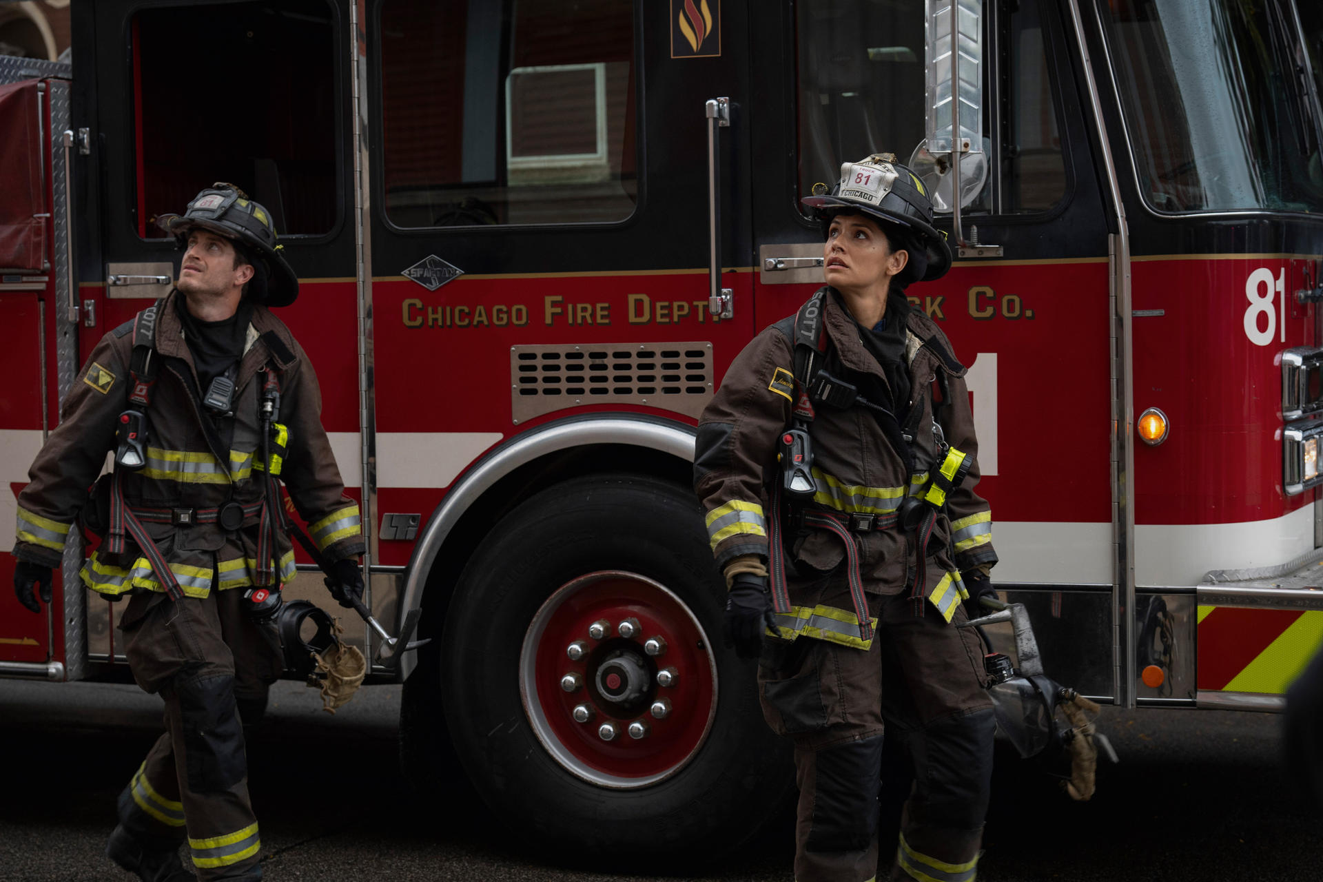 Zwei Feuerwehrmänner in Uniform stehen in der Nähe eines roten Feuerwehrautos. Beide blicken nach oben. Auf dem Wagen steht Chicago Fire Dept.