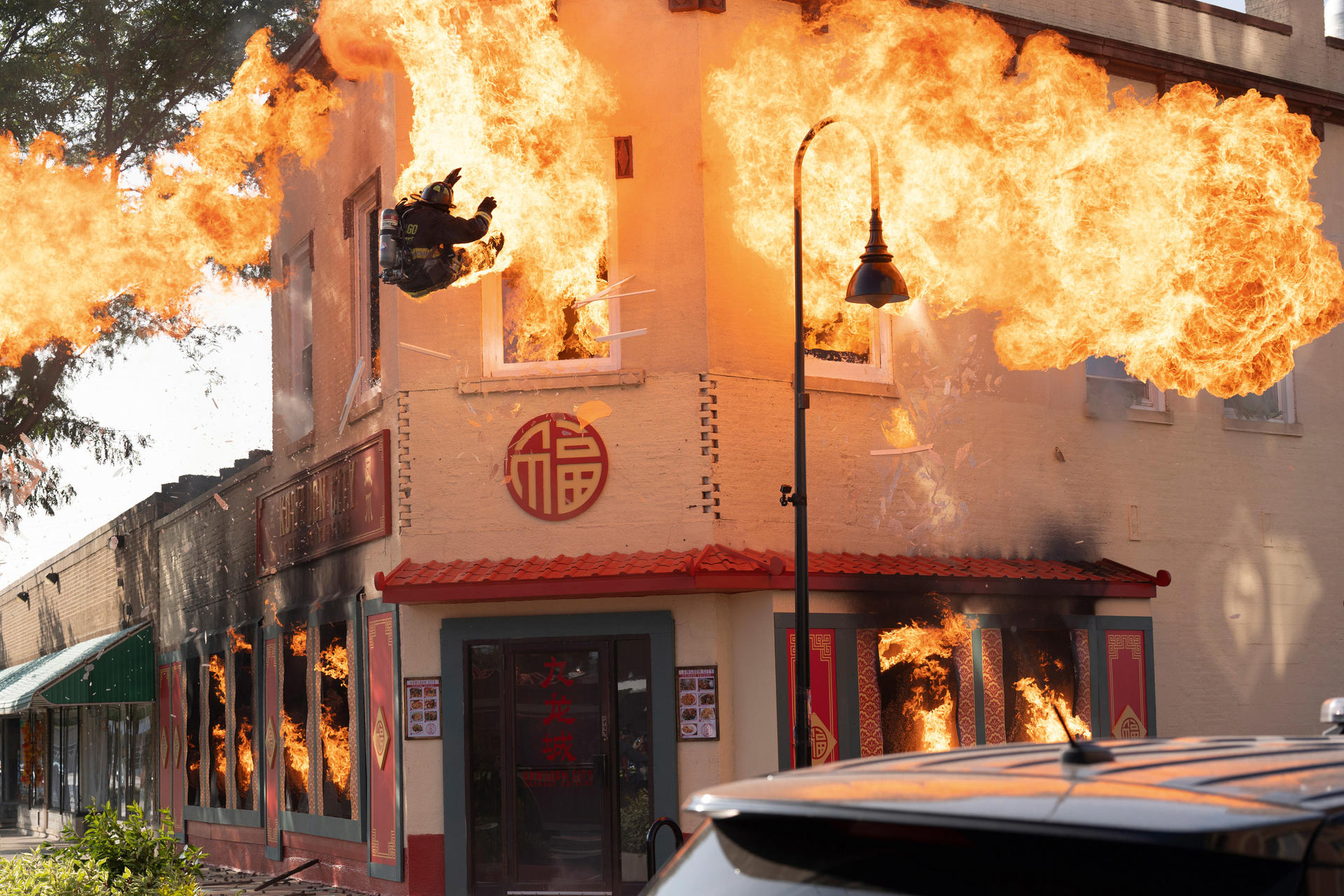 Ein Gebäude steht in Flammen. Ein Feuerwehrmann springt aus einem Fenster. Feuer breitet sich schnell aus. Eine Straßenlaterne steht.