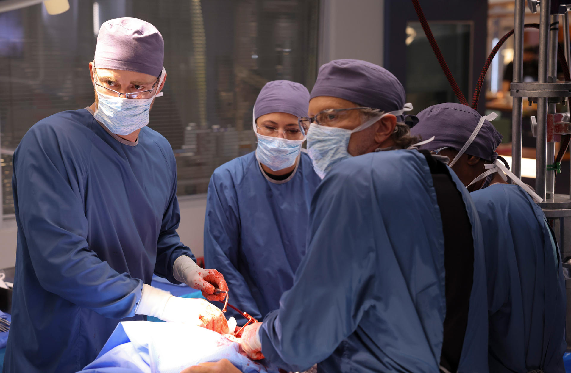 Ein OP-Team in blauen OP-Kitteln und -Mützen operiert einen Patienten. Blut ist sichtbar. Sie tragen Masken und Handschuhe.
