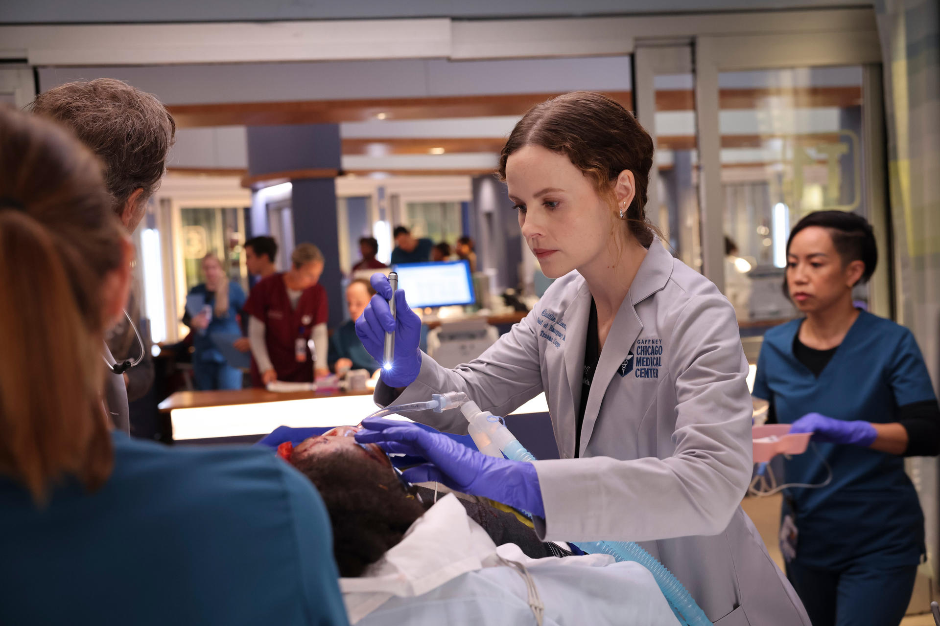 Ein Arzt in grauem Mantel und blauen Handschuhen untersucht einen Patienten in einer medizinischen Umgebung. Anderes medizinisches Personal ist anwesend.