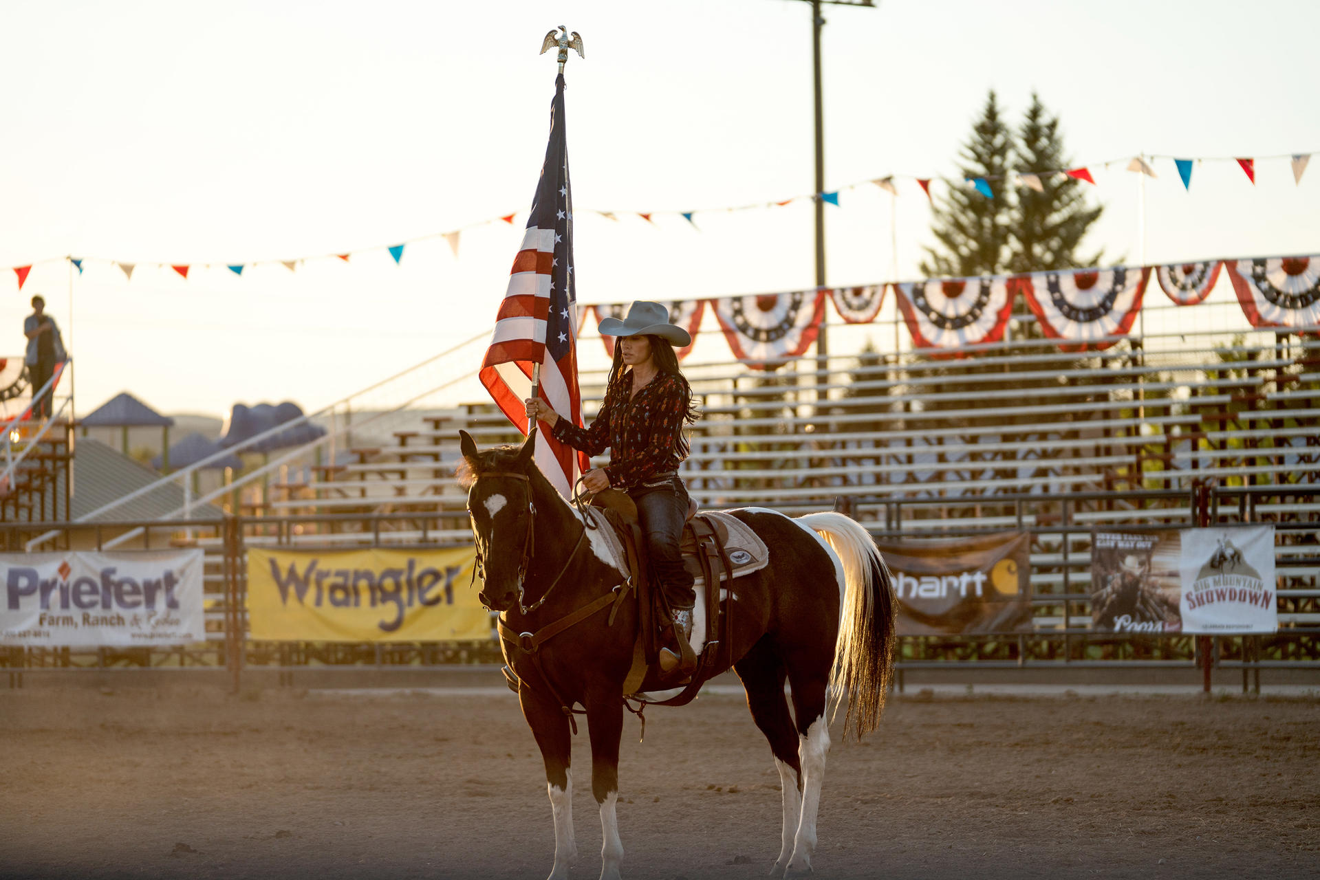 Eine Frau auf einem Pferd hält eine amerikanische Flagge. Rodeo-Arena mit Bannern und Zuschauern im Hintergrund. Abendlicht.