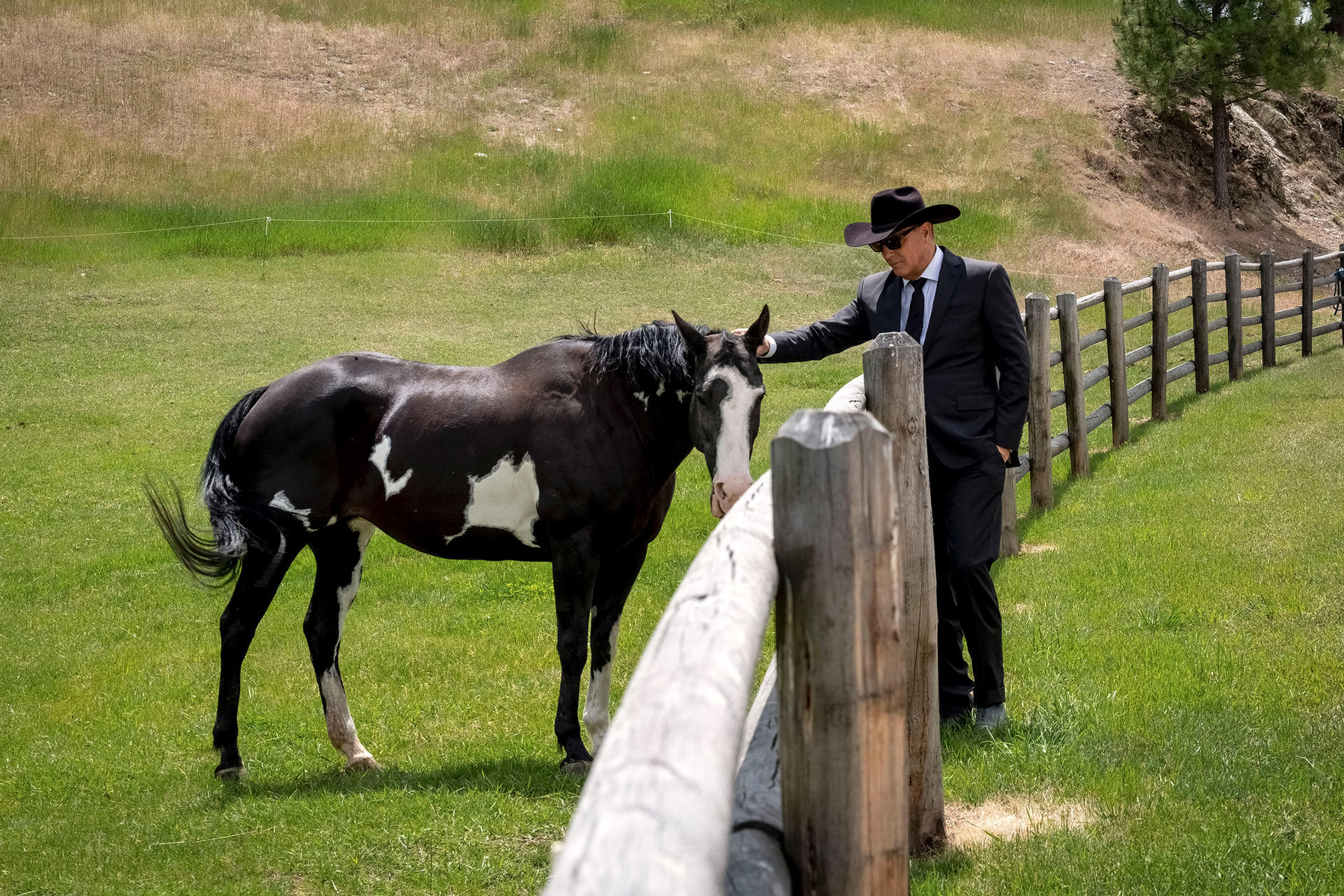 Ein Mann in Anzug und Hut streichelt ein schwarz-weißes Pferd auf einer Wiese neben einem Holzzaun.