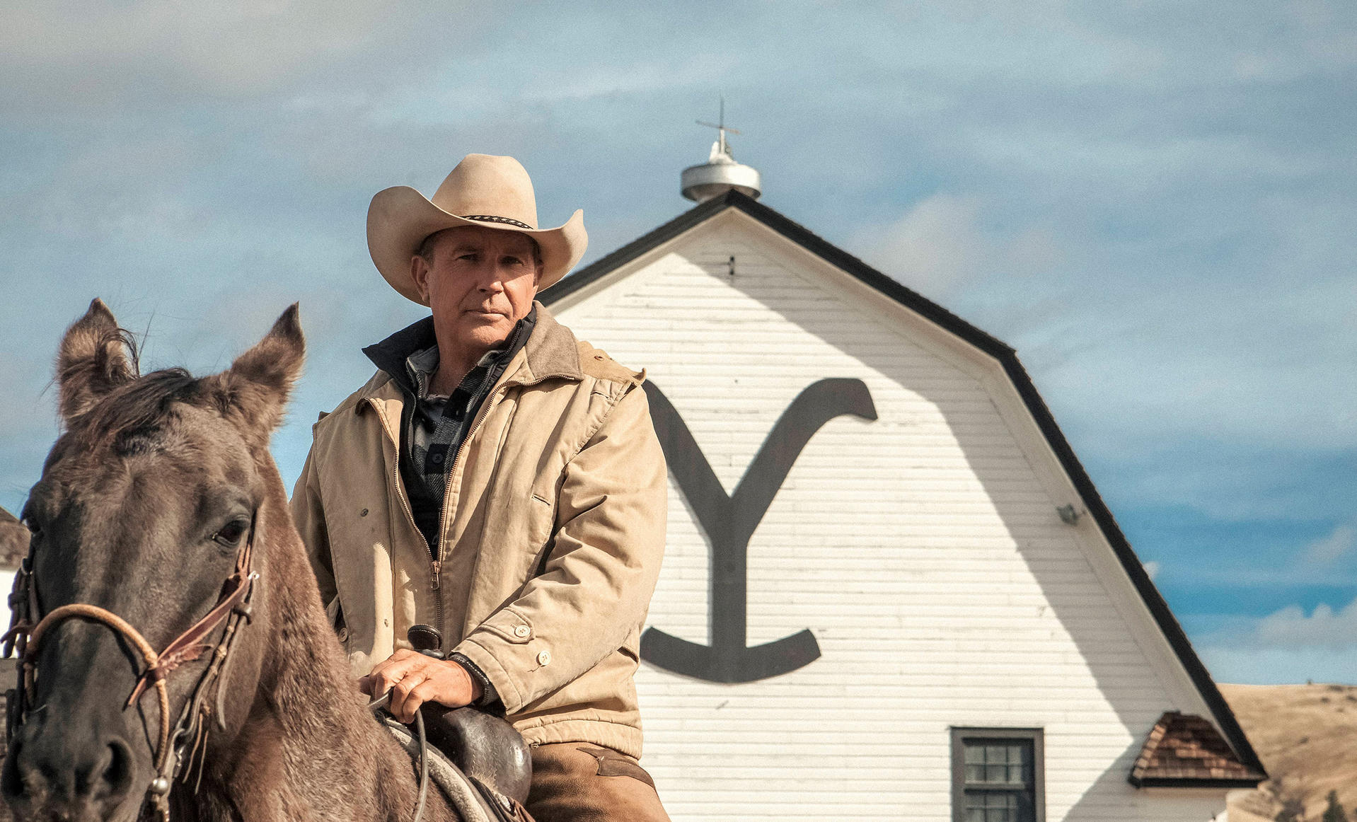 Ein Mann mit Cowboyhut auf einem braunen Pferd, eine weiße Scheune mit schwarzem "Y"-Logo im Hintergrund und ein klarer Himmel.