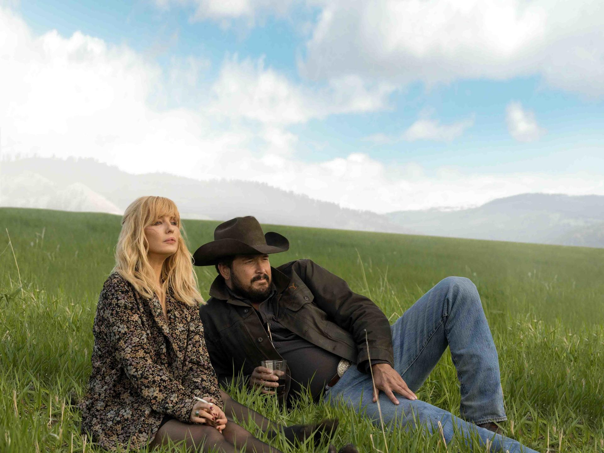 Ein Mann und eine Frau liegen im Gras. Die Frau hat blonde Haare und eine Blumenjacke. Der Mann trägt einen Hut und eine Jacke. Wolkenverhangener Himmel.