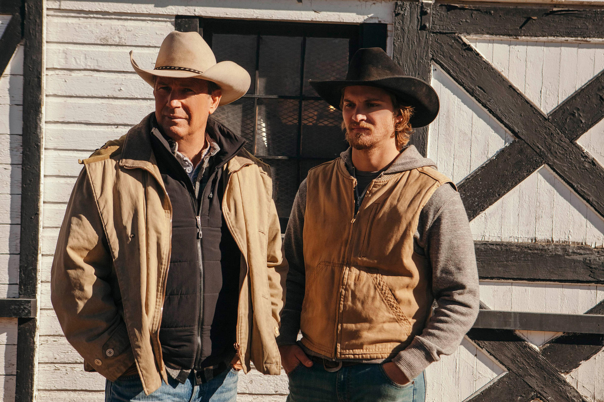 Zwei Männer in Cowboyhüten und Jacken stehen an einer weißen Holzwand mit schwarzen Akzenten. Einer hat eine beige Jacke, der andere eine braune.