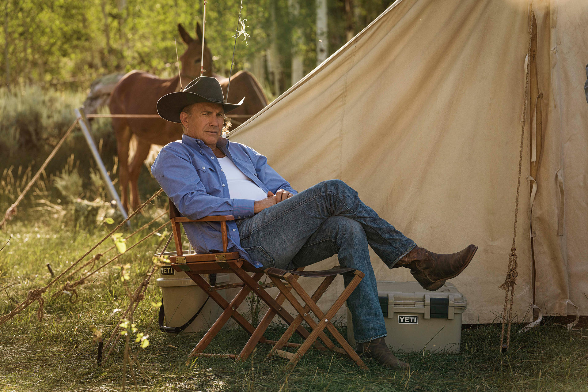 Ein Mann mit Cowboyhut sitzt in einem Stuhl. Ein Zelt und eine Kühlbox sind hinter ihm. Pferde sind im Hintergrund.