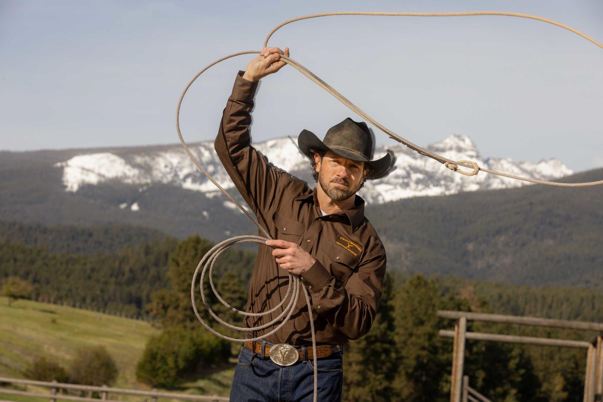 Ein Mann mit Cowboyhut und Hemd hält ein Seil vor dem Hintergrund von Bergen und Bäumen. Er wirft ein Lasso.