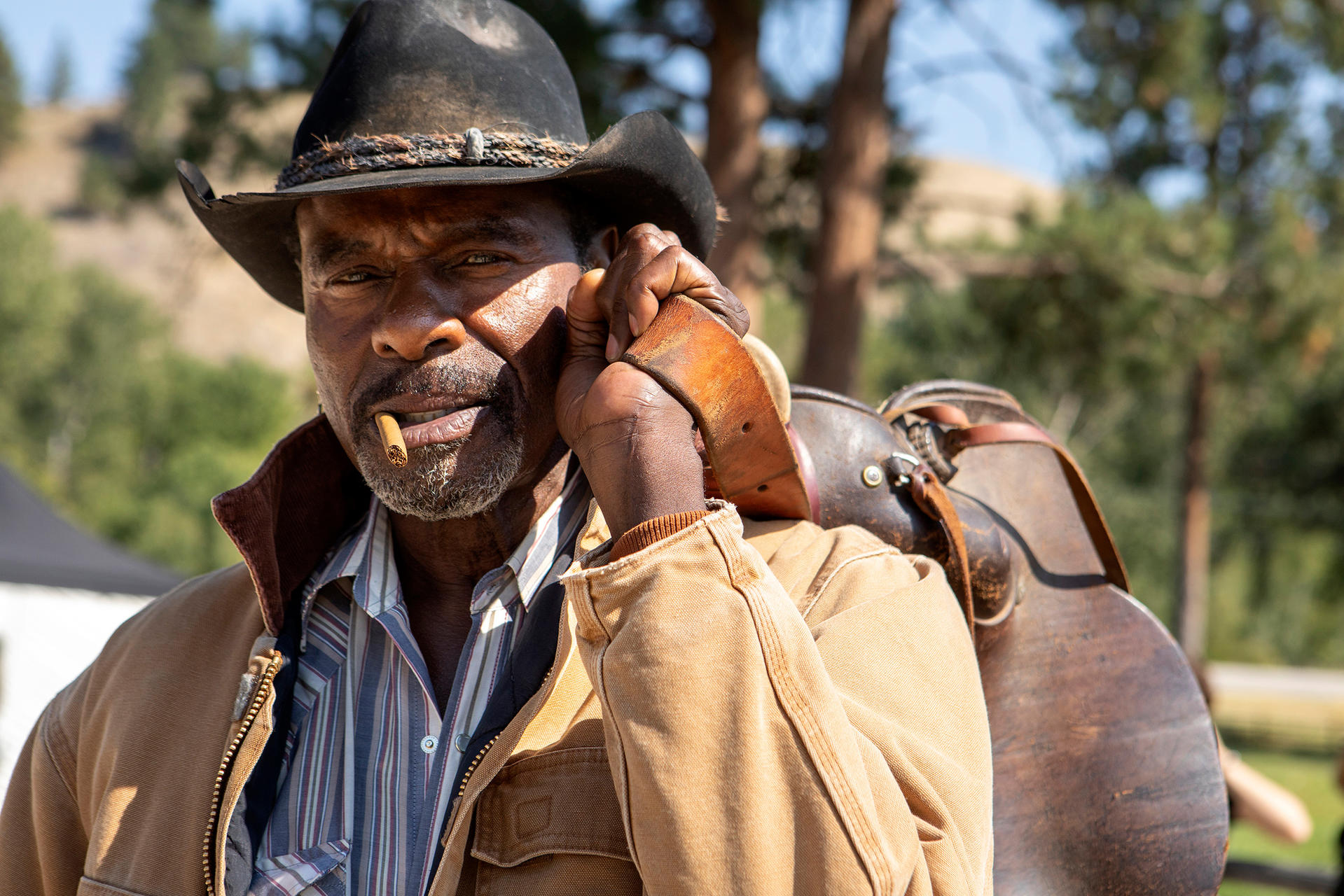 Ein älterer schwarzer Mann mit Cowboyhut hält einen Sattel. Er raucht eine Zigarre, trägt eine beigefarbene Jacke und steht vor Bäumen.