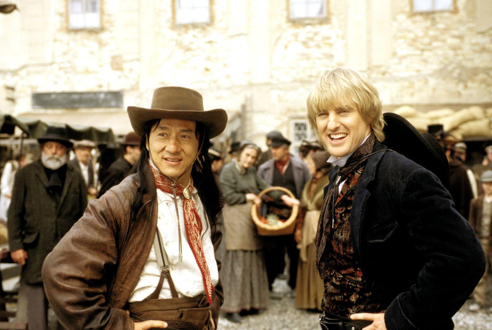 Zwei Männer in Kostümen lächeln in einem Stadtplatz. Einer trägt einen Cowboyhut, der andere einen Anzug.