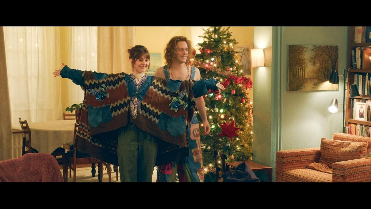 Zwei Personen mit Patchwork-Jacke, Arme ausgestreckt. Weihnachtsbaum im Hintergrund mit Lichtern. Gemütliches Zimmer mit warmer Beleuchtung.