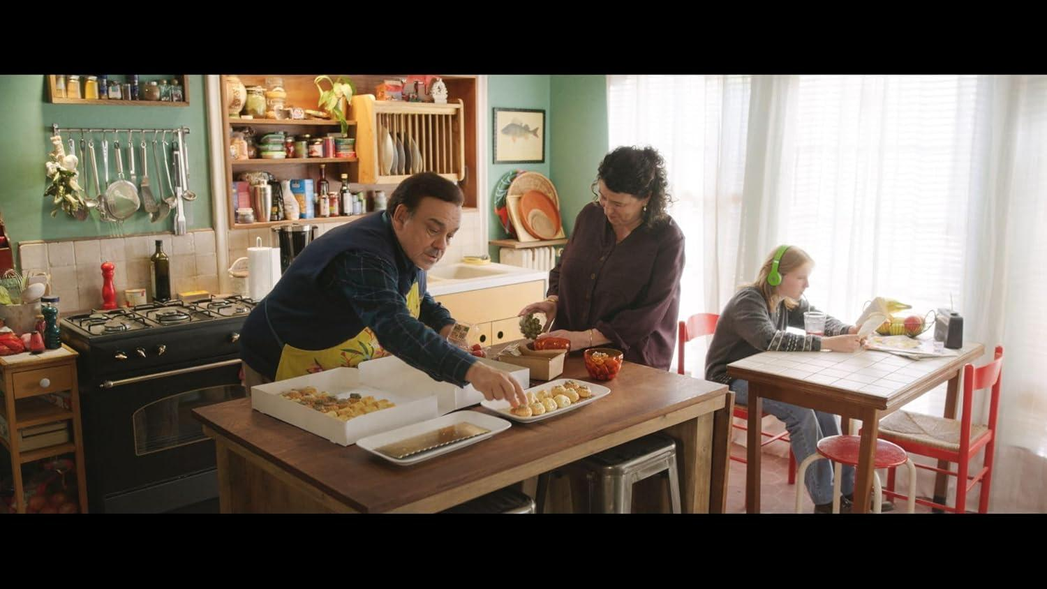 Eine Familie in einer Küche, mit einem Mann, der Gebäck anrichtet, einer Frau, die Essen zubereitet, und einem Mädchen mit Musik.