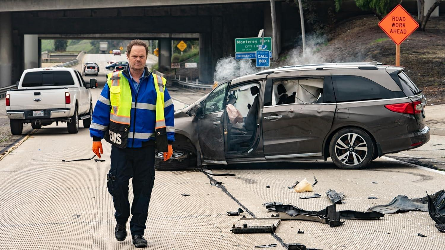 Unfallort. Ein Sanitäter steht auf der Straße. Beschädigter Minivan neben dem Arbeiter. Baustellenschild sichtbar.