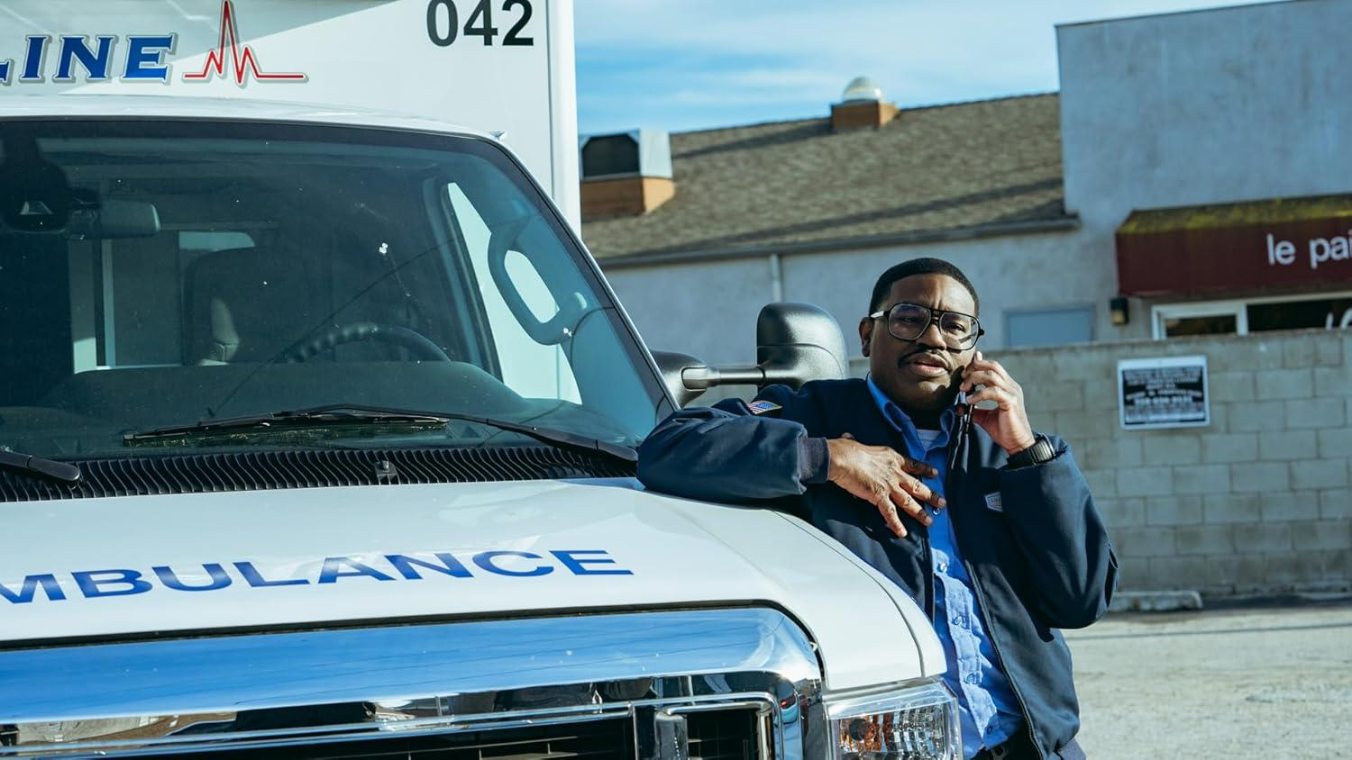 Ein Mann in Uniform lehnt an einem Krankenwagen und telefoniert. Im Hintergrund ein Gebäude mit Schild und blauer Himmel.