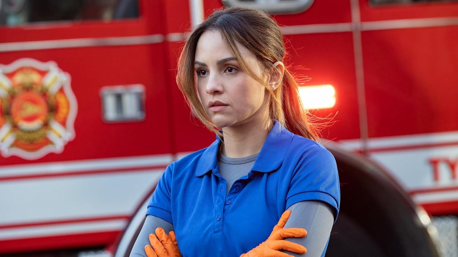Eine Frau in blauem Hemd und orangefarbenen Handschuhen steht vor einem roten Feuerwehrauto. Sie sieht besorgt aus.
