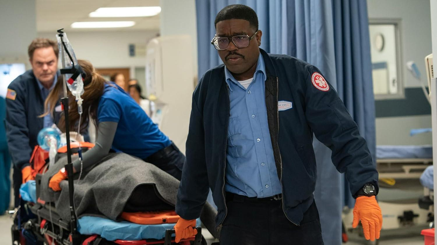 Ein Sanitäter mit Brille und Handschuhen schaut in die Kamera. Andere Sanitäter schieben einen Patienten auf einer Trage im Krankenhausgang.