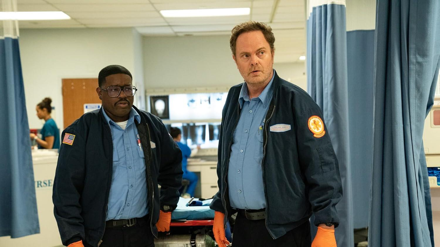 Zwei Männer in Sanitäteruniformen stehen in einem Krankenhausflur. Einer trägt eine Brille. Im Hintergrund ist eine Frau zu sehen.