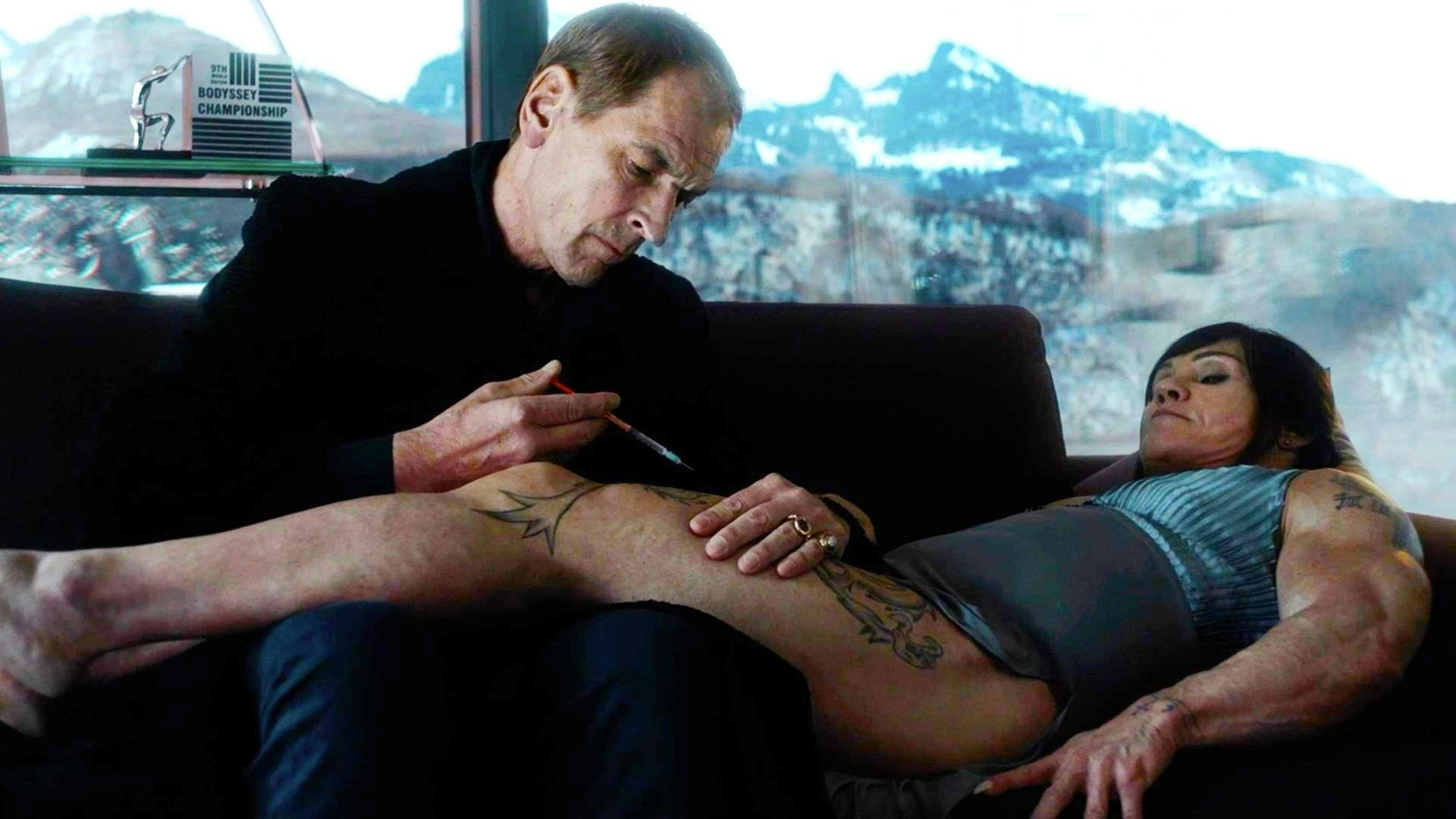Mann injiziert Bein einer Frau. Sie liegt auf einem Sofa, tätowiert. Trophäe im Hintergrund. Schneebedeckte Berge durch das Fenster sichtbar.