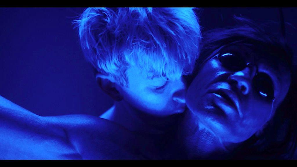 Zwei Personen in blauem Licht. Einer trägt eine Sonnenbrille, der andere hat blondes Haar. Sie sind nah beieinander, das Gesicht des einen berührt den Hals des anderen.