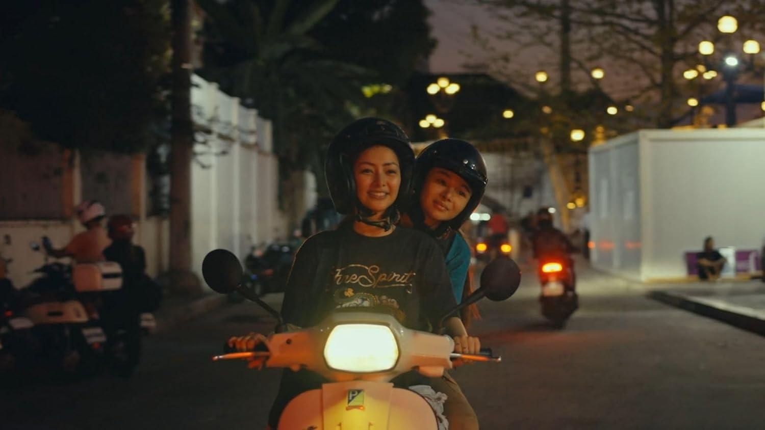 Zwei Frauen fahren nachts mit einem Roller. Sie tragen Helme. Im Hintergrund sind andere Roller und Lichter zu sehen.