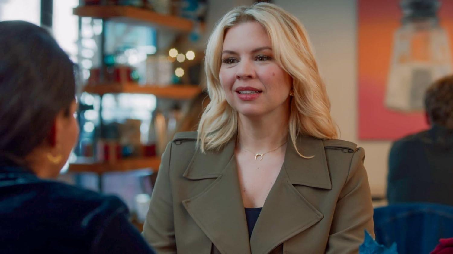 Eine blonde Frau in einem olivfarbenen Mantel lächelt. Sie trägt eine Halbmondkette. Eine weitere Person ist im Vorder- und Hintergrund.